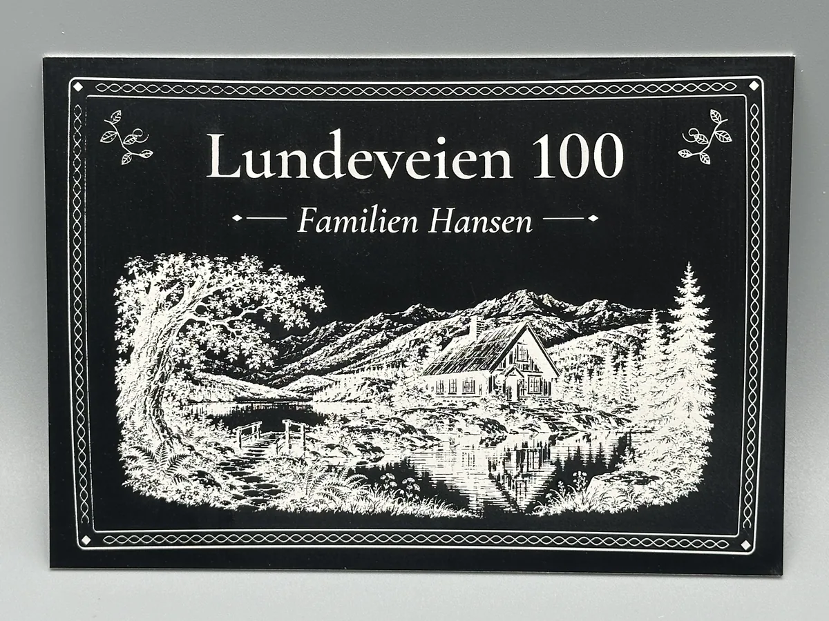 Sort aluminiumsskilt med ramme, adressen Lundeveien 100, «Familien Hansen» og et detaljert gravert motiv av en hytte ved et fjellvann