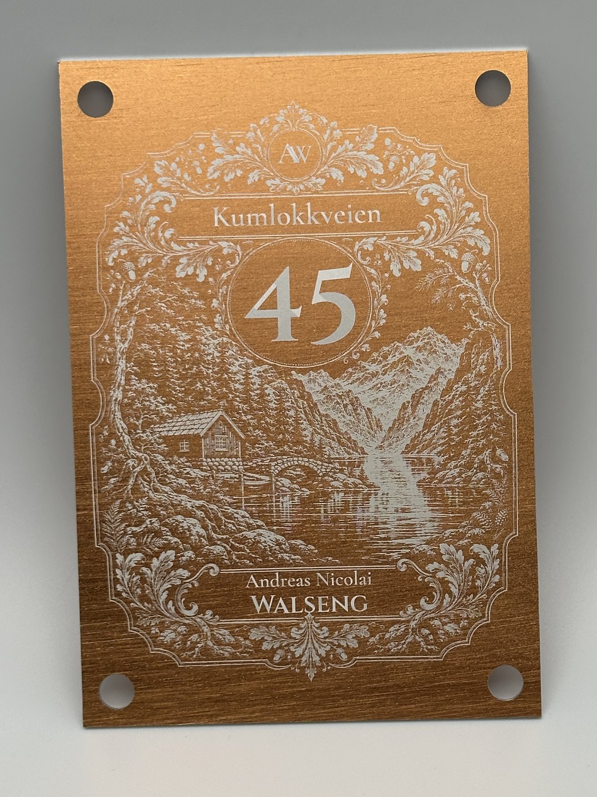 Husskilt i kobberfarget aluminium, 105 × 150 mm med fire hull: gravert ornamentramme, husnummer, gatenavn, navn og fjordmotiv