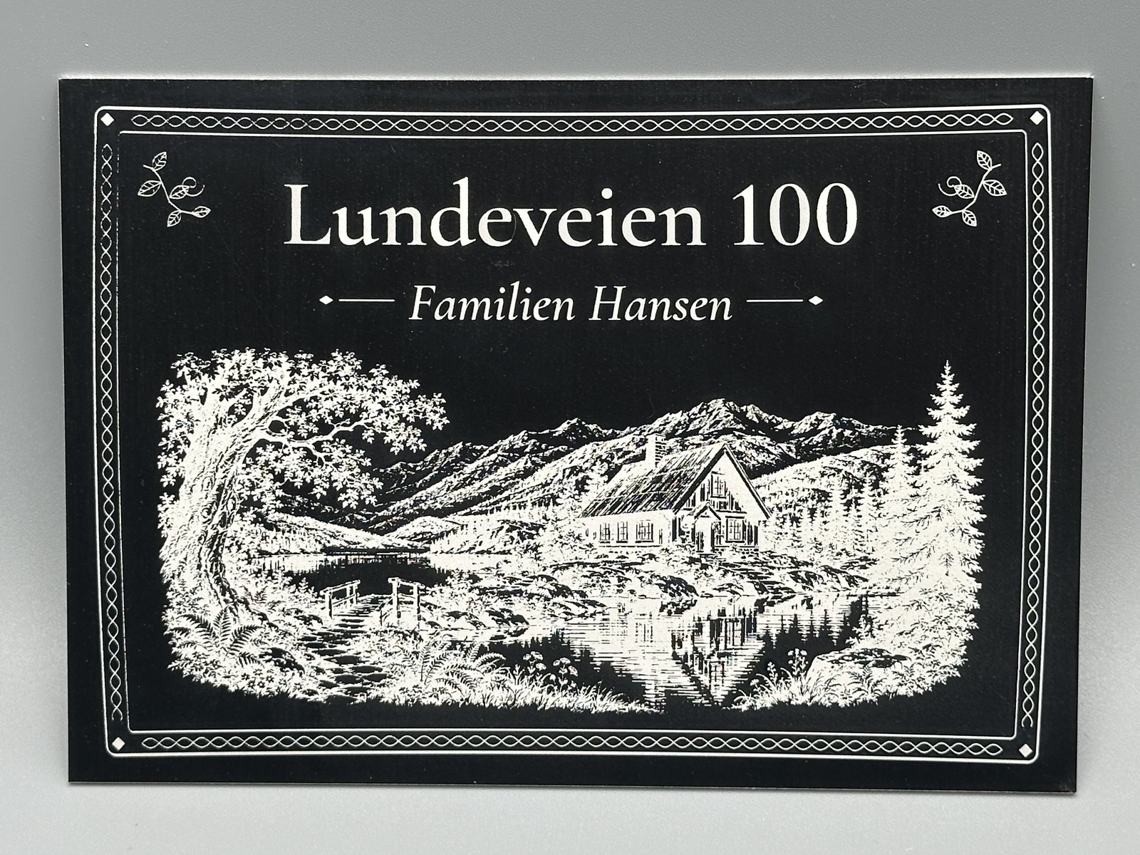 Dørskilt i sort aluminium med ramme, adressen Lundeveien 100, «Familien Hansen» og et gravert motiv av en hytte ved et fjellvann
